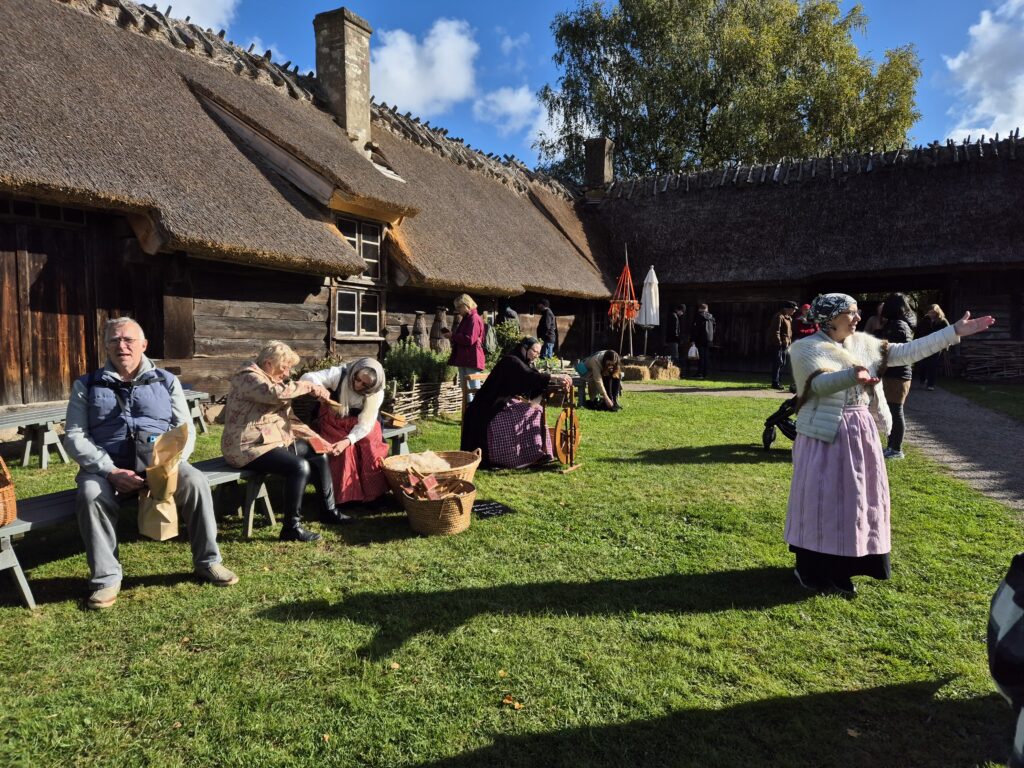 Utanför en gammal trästuga står en äldre dam och en dam sitter och gör egen ull.